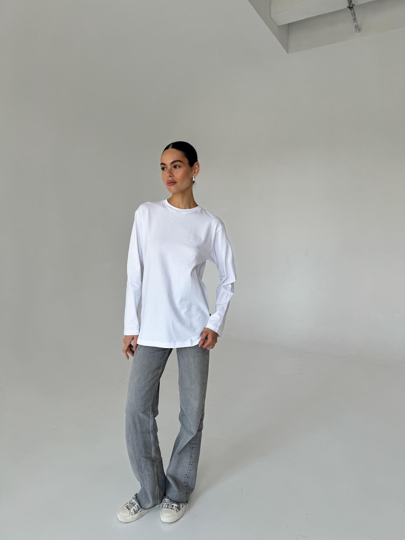 May include: A white long-sleeve shirt and gray jeans. The shirt has a crew neck and a relaxed fit. The jeans are a light gray wash with a flared leg. The outfit is paired with white sneakers.