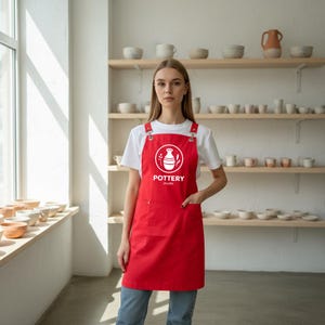 Może przedstawiać: Czerwony fartuch z napisem "Pottery Studio" i logo wazonu. Fartuch ma dwie przednie kieszenie i regulowane paski. Osoba ma na sobie białą koszulkę i niebieskie dżinsy.