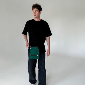 Puede incluir: Una camiseta negra, pantalones vaqueros oscuros y zapatillas blancas combinados con una bolsa utilitaria rectangular verde con correas de cuero negro y botones de presión metálicos plateados que se lleva en la cintura con dos correas horizontales.