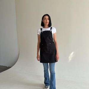 Pode incluir: Um avental preto com al&ccedil;as ajust&aacute;veis e bolsos. O avental &eacute; usado sobre uma camiseta branca e jeans azuis. A modelo est&aacute; usando t&ecirc;nis brancos.