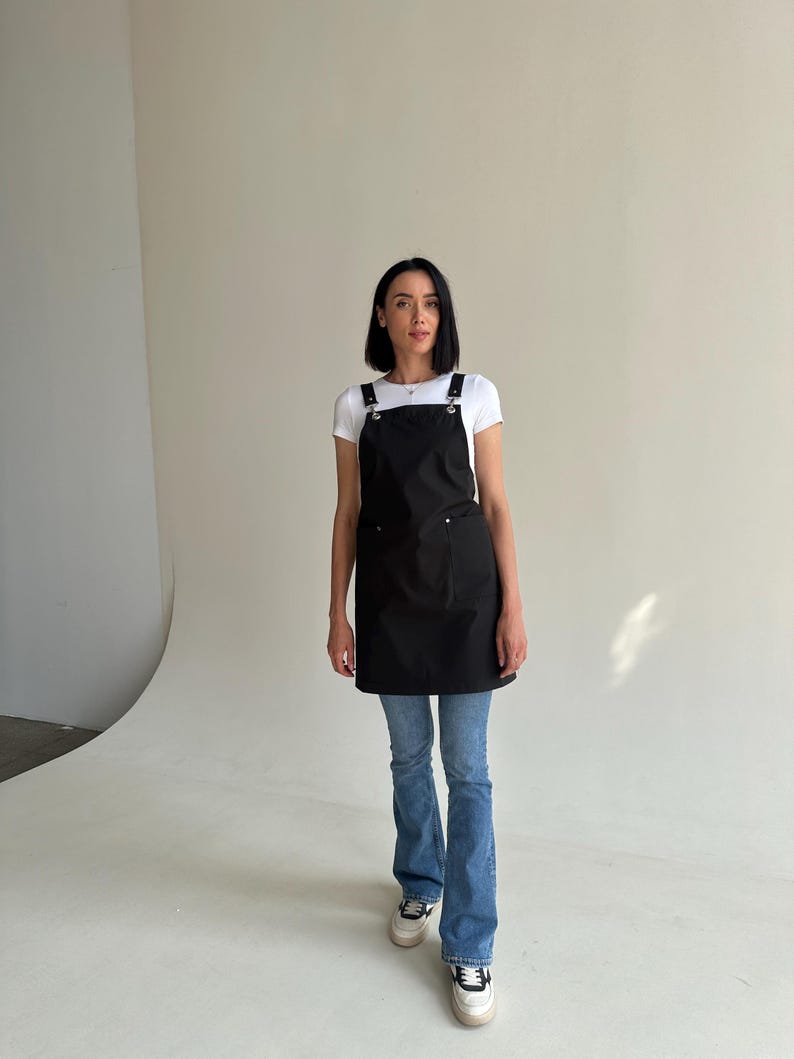 Pode incluir: Um avental preto com al&ccedil;as ajust&aacute;veis e bolsos. O avental &eacute; usado sobre uma camiseta branca e jeans azuis. A modelo est&aacute; usando t&ecirc;nis brancos.