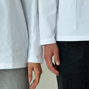 May include: Two people wearing white long-sleeve shirts and dark-colored pants. The hands of the two people are touching. The image is a medium shot, with a neutral background.