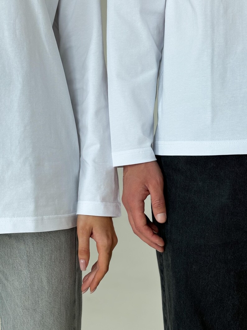 May include: Two people wearing white long-sleeve shirts and dark-colored pants. The hands of the two people are touching. The image is a medium shot, with a neutral background.