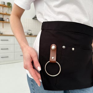 Può includere: Grembiule nero in vita con un'ampia tasca e un passante in pelle marrone con un anello argentato. Il grembiule ha due rivetti argentati e una piccola tasca. Indossato sopra una maglietta bianca e jeans blu.