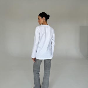 May include: A person wearing a white long-sleeve shirt and gray jeans. The jeans have a flared leg design. The person is also wearing white sneakers.