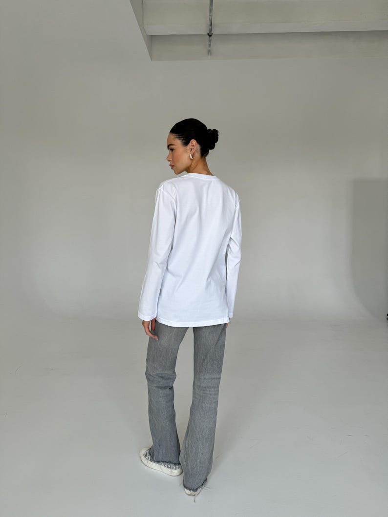 May include: A person wearing a white long-sleeve shirt and gray jeans. The jeans have a flared leg design. The person is also wearing white sneakers.