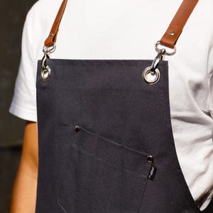 May include: A dark gray canvas apron featuring brown leather straps attached with silver-toned metal lobster clasps and grommets. The apron includes a front pocket secured with silver rivets and a small black fabric tag labeled with the text PROFIT.