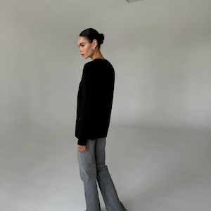May include: A person wearing a black long-sleeve shirt and gray flared jeans. The person is wearing white sneakers. The background is a plain white studio setting.