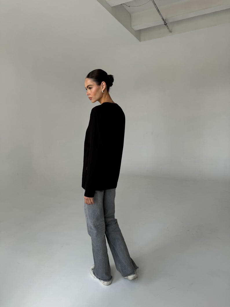May include: A person wearing a black long-sleeve shirt and gray flared jeans. The person is wearing white sneakers. The background is a plain white studio setting.