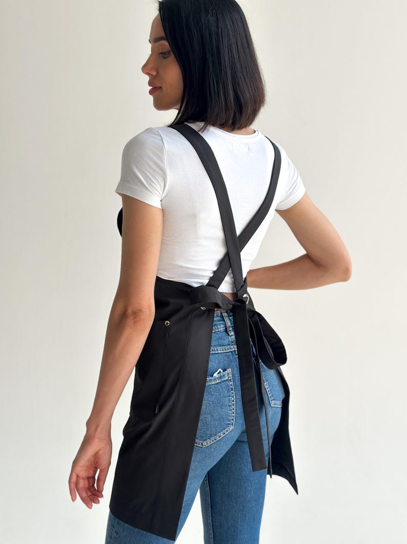 Pode incluir: Avental preto com al&ccedil;as cruzadas nas costas e cinto para amarrar. O avental &eacute; feito de um tecido escuro e liso e possui um bolso na frente. O avental &eacute; usado sobre uma camiseta branca e jeans azul.