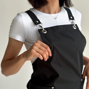 Puede incluir: Delantal negro con correas ajustables y herrajes plateados. El delantal tiene dos bolsillos delanteros y un diseño tipo peto. Las correas se sujetan con anillas y cierres metálicos. Se lleva sobre una camiseta blanca.