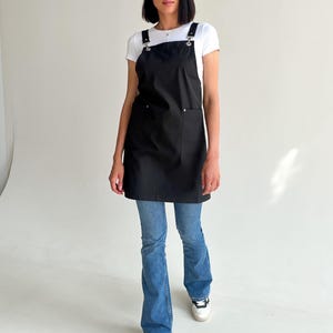 Pode incluir: Um avental preto com um grande bolso frontal e al&ccedil;as ajust&aacute;veis. O avental &eacute; usado sobre uma camiseta branca e jeans azuis. As al&ccedil;as s&atilde;o presas com ferragens de metal prateado.