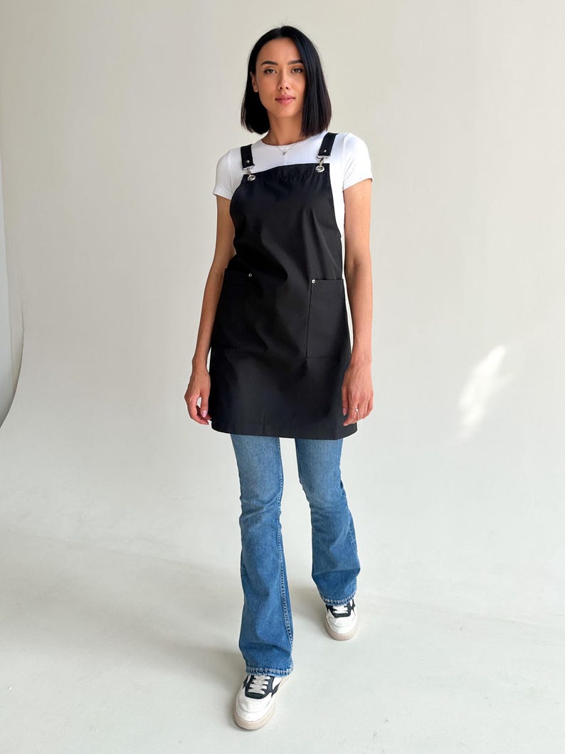 Pode incluir: Um avental preto com um grande bolso frontal e al&ccedil;as ajust&aacute;veis. O avental &eacute; usado sobre uma camiseta branca e jeans azuis. As al&ccedil;as s&atilde;o presas com ferragens de metal prateado.