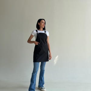 Pode incluir: Um avental preto com al&ccedil;as ajust&aacute;veis e ferragens met&aacute;licas. O avental tem um bolso frontal e &eacute; usado sobre uma camiseta branca e jeans azuis. A modelo est&aacute; usando t&ecirc;nis brancos.