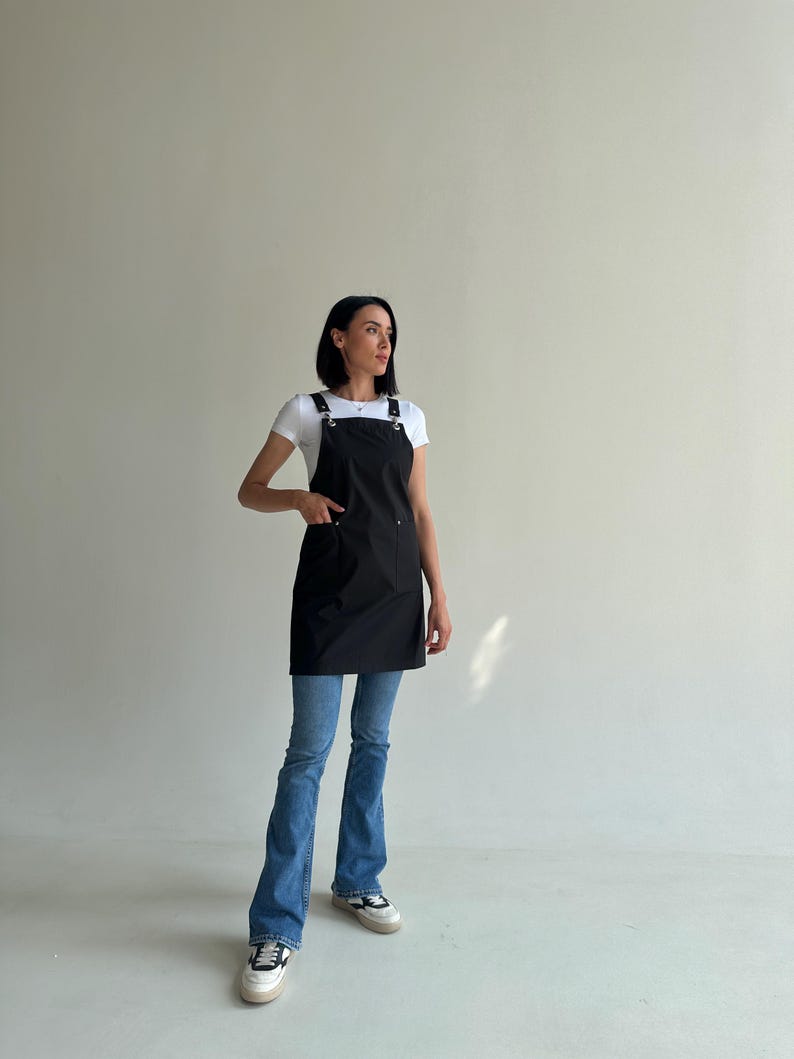 Pode incluir: Um avental preto com al&ccedil;as ajust&aacute;veis e ferragens met&aacute;licas. O avental tem um bolso frontal e &eacute; usado sobre uma camiseta branca e jeans azuis. A modelo est&aacute; usando t&ecirc;nis brancos.