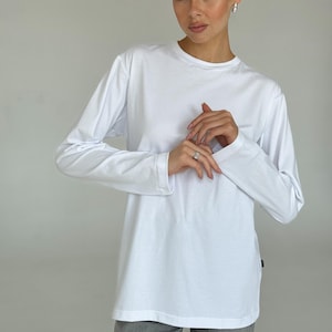 May include: A white long-sleeve shirt with a crew neck. The shirt is a solid color and appears to be made of a soft material. The model is wearing gray pants.