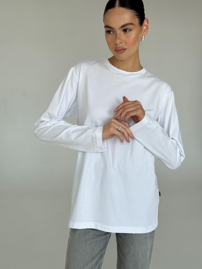 May include: A white long-sleeve shirt with a crew neck. The shirt is a solid color and appears to be made of a soft material. The model is wearing gray pants.