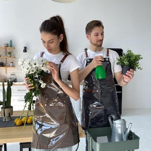 May include: Two people wearing clear vinyl aprons with brown leather straps. One holds white flowers, the other a potted plant and a green spray bottle. A green rolling cart with a watering can and other items is in the foreground.