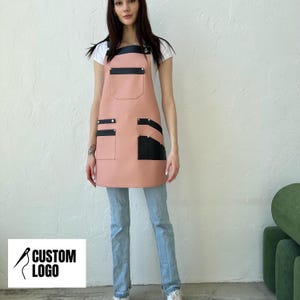 May include: A light pink apron featuring black leather-like trim and pockets. The apron has a chest pocket, two lower pockets, and silver-toned metal rivets securing the straps and pocket corners. It is worn over a white t-shirt and blue jeans.