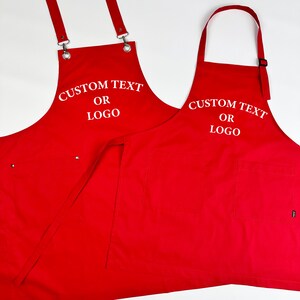 Może przedstawiać: Dwa czerwone fartuchy z regulowanymi paskami i kieszeniami. Na każdym fartuchu znajduje się biały napis "CUSTOM TEXT OR LOGO". Fartuchy wykonane są z wytrzymałej tkaniny i przeznaczone do użytku w kuchni lub rzemiośle.