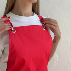 May include: A bright red apron with silver metal hardware. The apron has adjustable straps with large metal rings and is worn over a white t-shirt. The apron is made of a smooth, solid fabric.