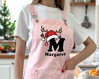Op de afbeelding: Een lichtroze schort met een kerstthema. Het schort heeft een rode kerstmuts met geweien, de letter "M" en de naam "Margaret" in het zwart. Het heeft twee zakken en verstelbare bandjes.