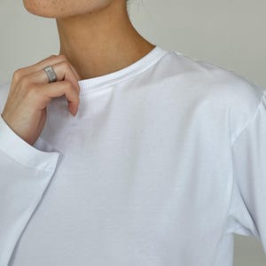 May include: A white long-sleeve shirt with a crew neck. The image also shows a silver ring and silver hoop earrings. The shirt is made of a soft, smooth fabric.