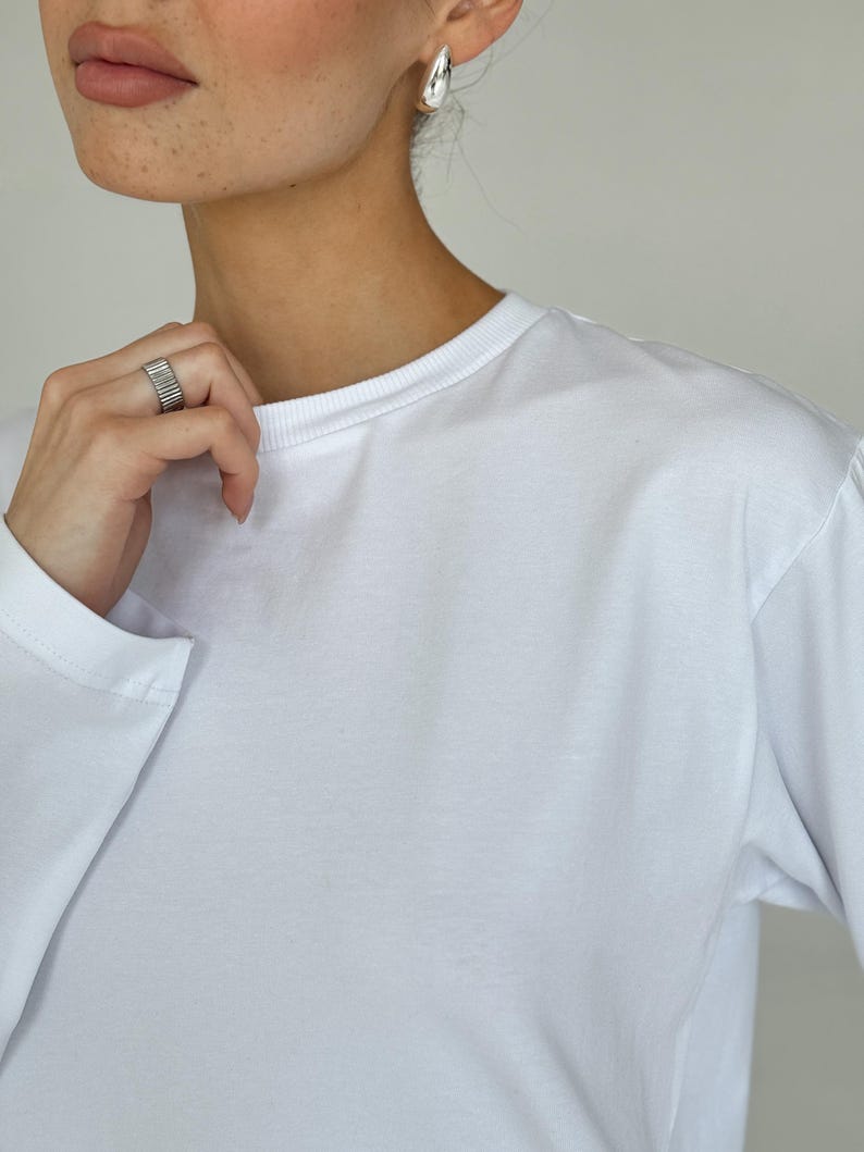 May include: A white long-sleeve shirt with a crew neck. The image also shows a silver ring and silver hoop earrings. The shirt is made of a soft, smooth fabric.
