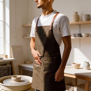May include: A dark olive green apron featuring brown leather straps secured with silver-toned metal rivets. The apron includes a chest pocket and two lower pockets, worn over a plain white short-sleeved t-shirt in a bright, sunlit studio setting.