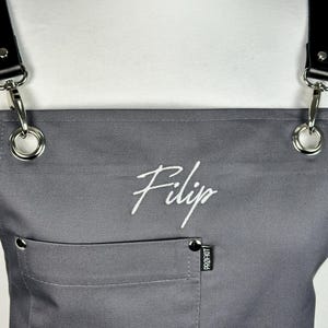 May include: Gray apron with black leather straps and silver hardware. The apron has a front pocket and the name "Filip" embroidered in white script. The tag on the pocket reads "PROF.KIT".