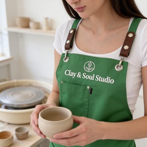 May include: A green apron featuring the white text Clay & Soul Studio and a circular logo. The apron has brown leather shoulder straps secured with silver-toned metal rivets and rings, and a front pocket with a small black fabric label attached.
