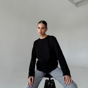 May include: A black long-sleeve shirt and gray jeans. The model is sitting on a black stool with a round seat and three legs. The background is a plain white wall.