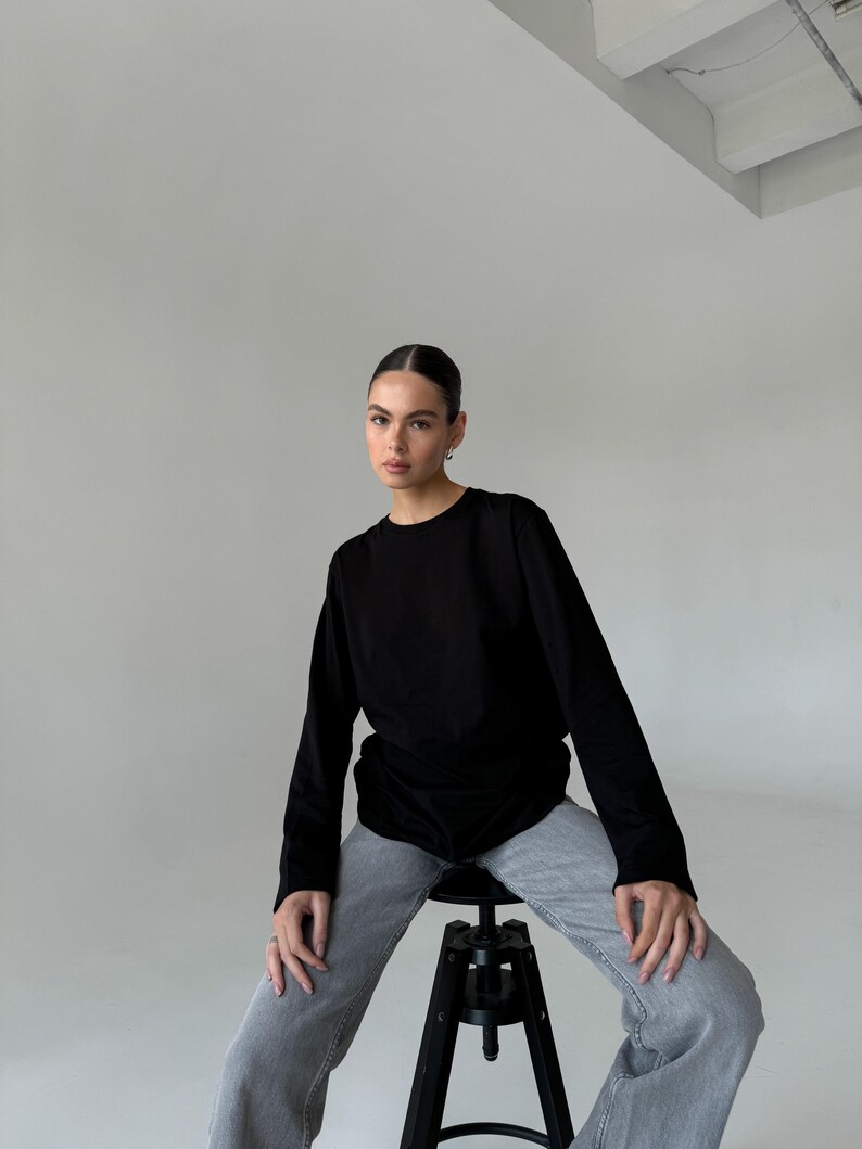 May include: A black long-sleeve shirt and gray jeans. The model is sitting on a black stool with a round seat and three legs. The background is a plain white wall.