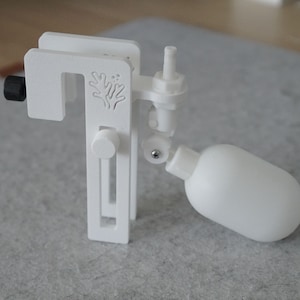 Könnte beinhalten: Ein weißes Kunststoff-Aquarium-Schwimmerventil mit Klemme, zylindrischem Körper und rundem Schwimmer. Die Klemme hat einen schwarzen Einstellknopf und ein dekoratives korallenartiges Design. Das Gerät dient zur Regulierung des Wasserstands.