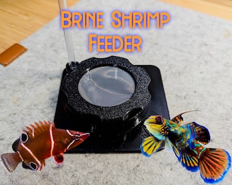May include: Black brine shrimp feeder with a magnifying glass, and the text "BRINE SHRIMP FEEDER". Two colorful fish are shown near the feeder. The feeder is on a gray surface.