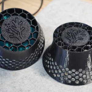 以下が含まれることがあります： ハニカム模様と木のようなエンブレムが付いた、黒色の3Dプリントスピーカーカバーが2つ。1つは大きく丸く、もう1つは円錐形です。大きい方のカバーの内側はティール色です。