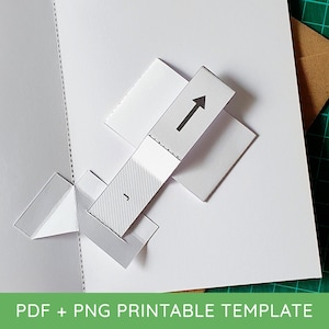 May include: A white paper pop-up card template featuring a central rectangular tab with a black arrow pointing upward and the letter J printed below it. The design includes perforated fold lines and geometric cutouts for assembly into a 3D structure.