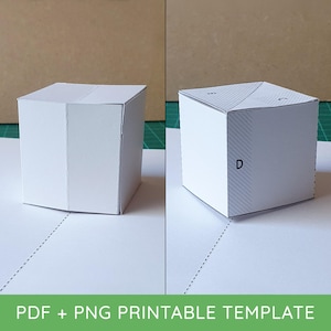 May include: A white paper cube craft template shown in two stages of assembly. The left cube is plain white, while the right cube features printed diagonal lines and the letters D, E, and C on its top and side panels, indicating folding lines.