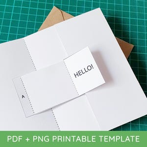 May include: A white paper craft template featuring a fold-out section with the text HELLO! printed in black. The template includes a section marked with the letter A and dashed lines for folding, positioned over a brown envelope on a green surface.