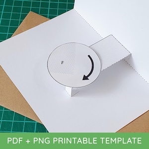 May include: A white paper pop-up card template featuring a circular rotating element with a black arrow and the letter F printed on it. The design includes dashed cut lines and a rectangular tab, presented as a printable PDF and PNG file format.