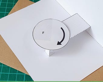 May include: A white paper pop-up card template featuring a circular rotating element with a black arrow and the letter F printed on it. The design includes dashed cut lines and a rectangular tab, presented as a printable PDF and PNG file format.