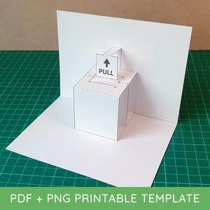Op de afbeelding: Een witte papieren pop-upkaart met een kubusontwerp. De kubus heeft een sleuf aan de bovenkant met een klein papieren lipje met de tekst "PULL" met een pijl. De kaart is open en onthult het pop-upelement. De onderkant van de kaart heeft de tekst "PDF + PNG PRINTABLE TEMPLATE".