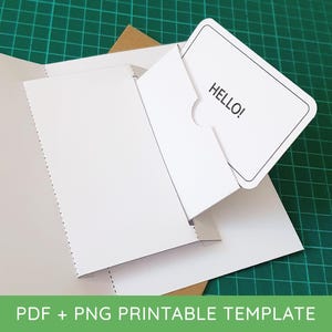 Peut inclure: Un modèle de porte-carte en papier blanc avec une pochette dotée d'une encoche arrondie. Une carte rectangulaire blanche aux coins arrondis avec le texte noir HELLO! est insérée dans la pochette. Le modèle est partiellement plié en papier cartonné.