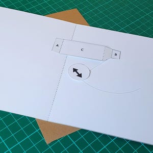 May include: A white paper template featuring a rectangular section labelled A, C, and B with diagonal line patterns. A circular white cutout with a black double-headed arrow icon is positioned below the rectangle, connected by a thin curved line.