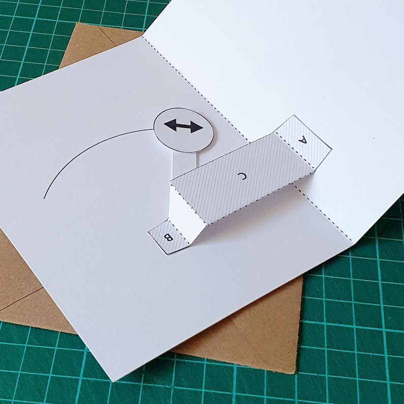 May include: A white paper pop-up card template featuring a three-dimensional bridge structure labelled with the letters A, B, and C. The design includes a circular icon with a double-headed arrow and a curved line, with dashed fold lines.