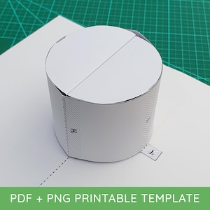 May include: A white paper craft template shaped into a cylinder with a flat circular top. The structure features printed assembly markings, including dashed fold lines and tabs labelled with the letters H1 and J2, designed for cutting and folding.