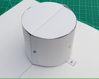 May include: A white paper craft template shaped into a cylinder with a flat circular top. The structure features printed assembly markings, including dashed fold lines and tabs labeled with the letters H1 and J2, designed for cutting and folding.