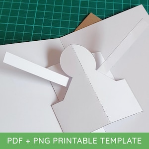 May include: A white paper template with cut-out shapes and dotted lines, partially folded. The image includes the text "PDF + PNG PRINTABLE TEMPLATE" in a green banner at the bottom. The background is a green cutting mat.