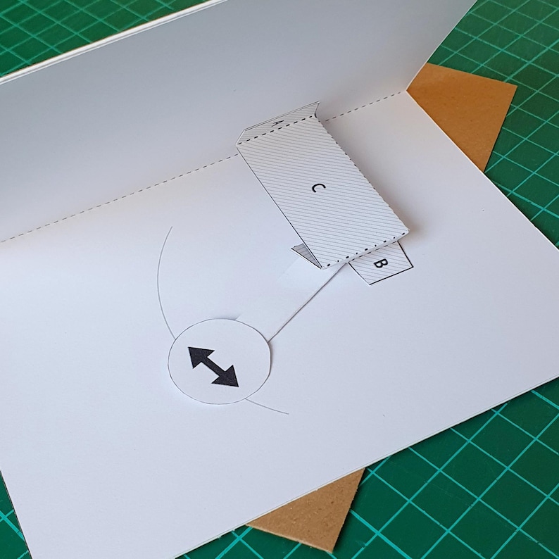 May include: A white paper pop-up card template featuring a rectangular section labelled with the letter C, a smaller tab labelled B, and a circular tab labelled with a black double-headed arrow icon. The card has dashed fold lines and printed guides.