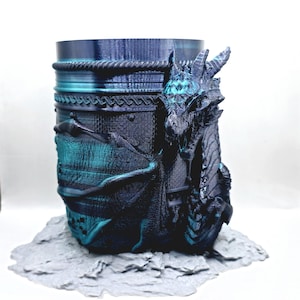 Peut inclure: Une tasse en forme de dragon imprimée en 3D avec un schéma de couleurs bleu et vert. Le dragon est enroulé autour de la tasse et présente un design détaillé. La tasse est posée sur une base grise.